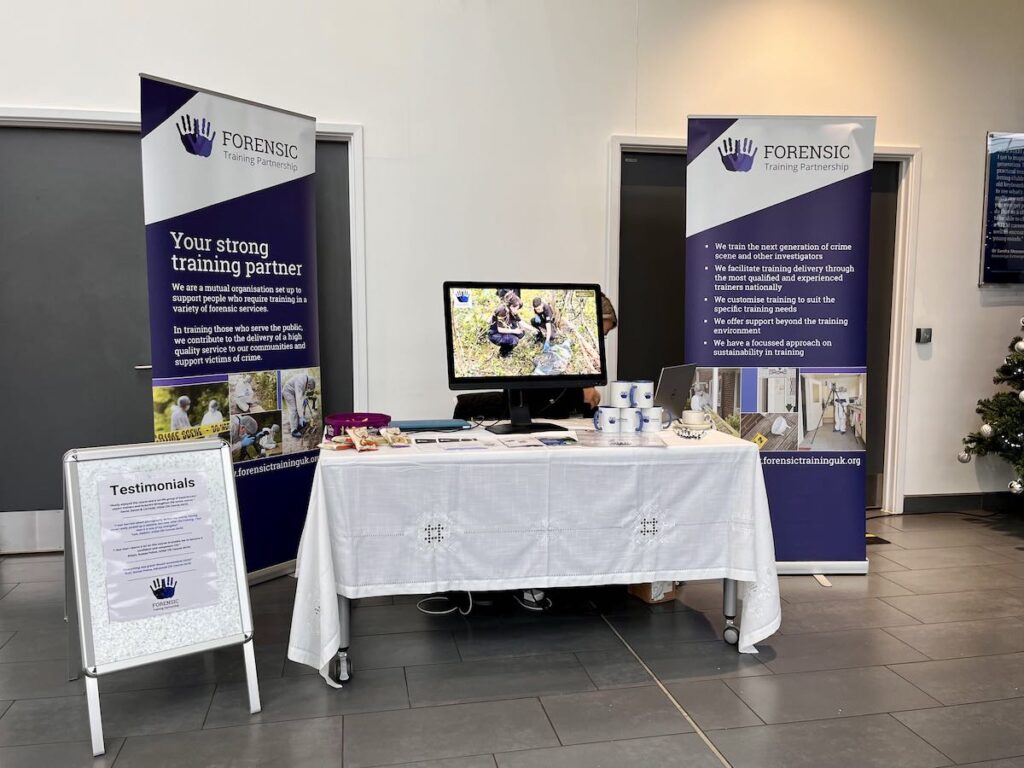 Supporting Forensic Professionals: Our Sponsorship of Key UK Conferences 1 A table with a white cloth and various items from The Forensic Training Partnership promoting their training courses.  with two banner stands either side of the table promoting FTP © MB/FTP 2025